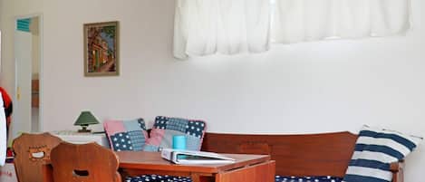 Una habitación acogedora con un banco de madera, una mesa y una silla. Se aprecia una ventana con cortinas blancas y un cuadro en la pared.