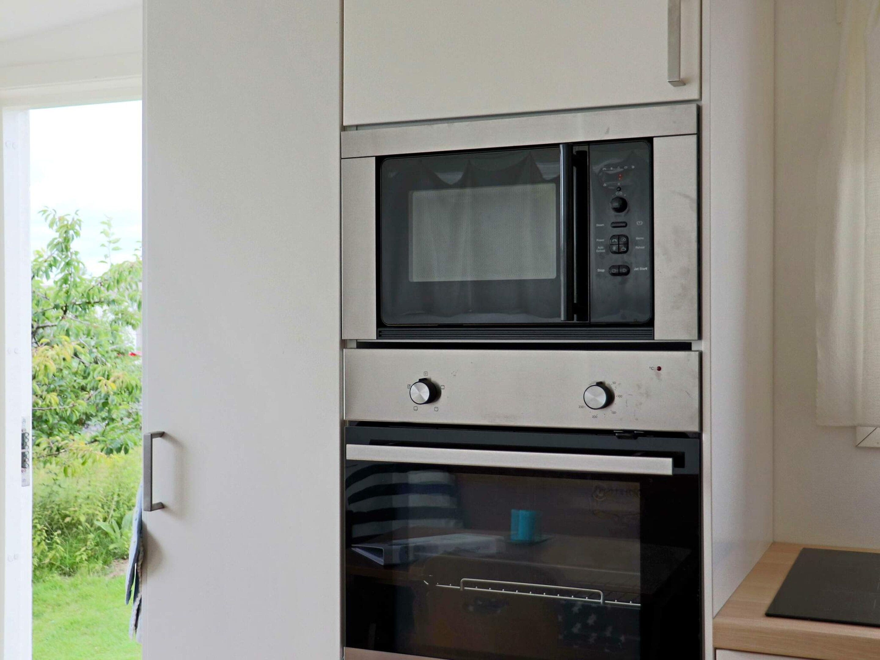 A kitchen with a built-in microwave and oven, a door with a handle, and a view of greenery outside.