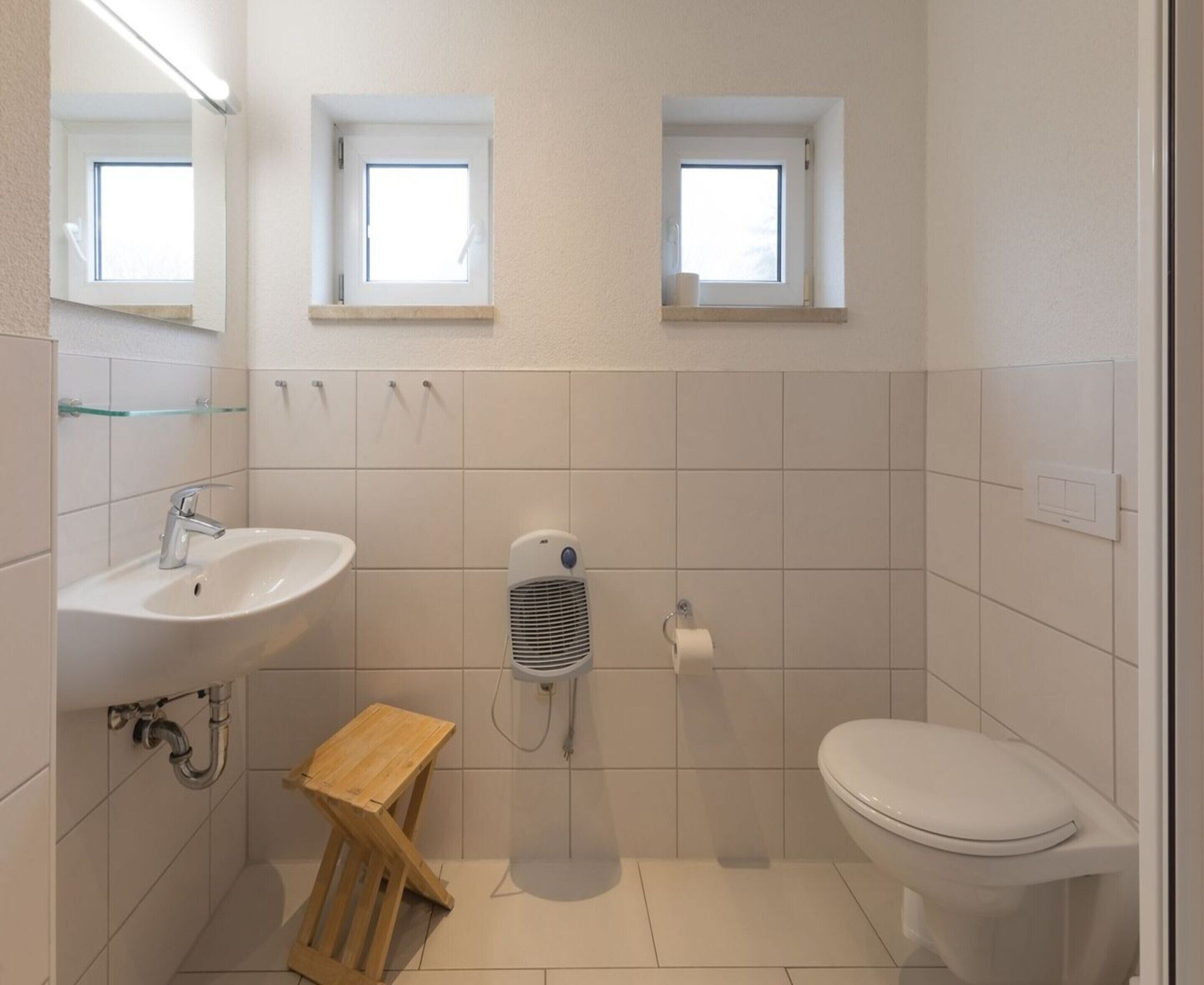 Une salle de bain moderne équipée d’un lavabo, de toilettes et d’une zone douche. Deux fenêtres laissent entrer la lumière naturelle.