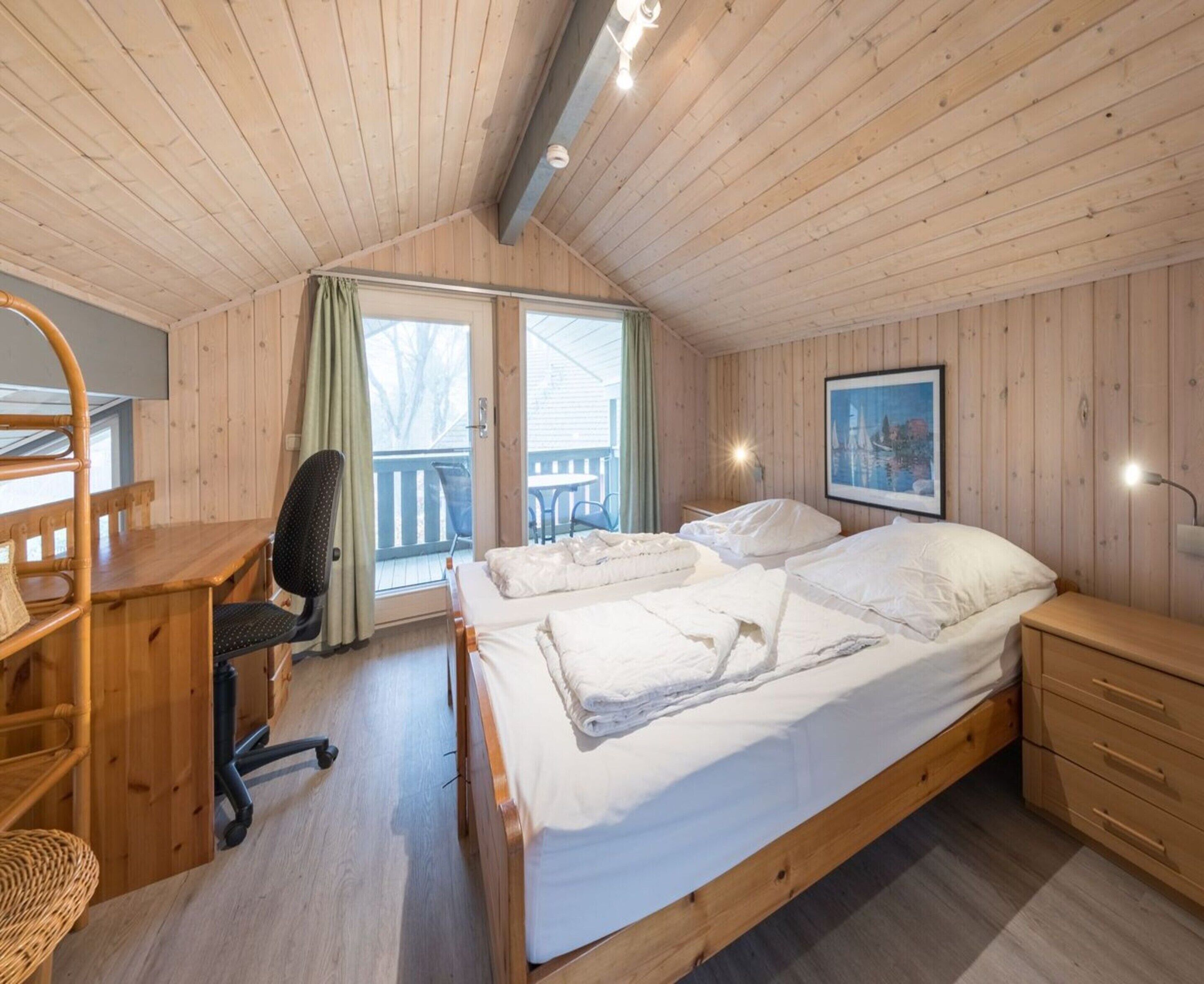 A wooden cabin bedroom with a bed, desk, chair, and a balcony with a table and chairs.