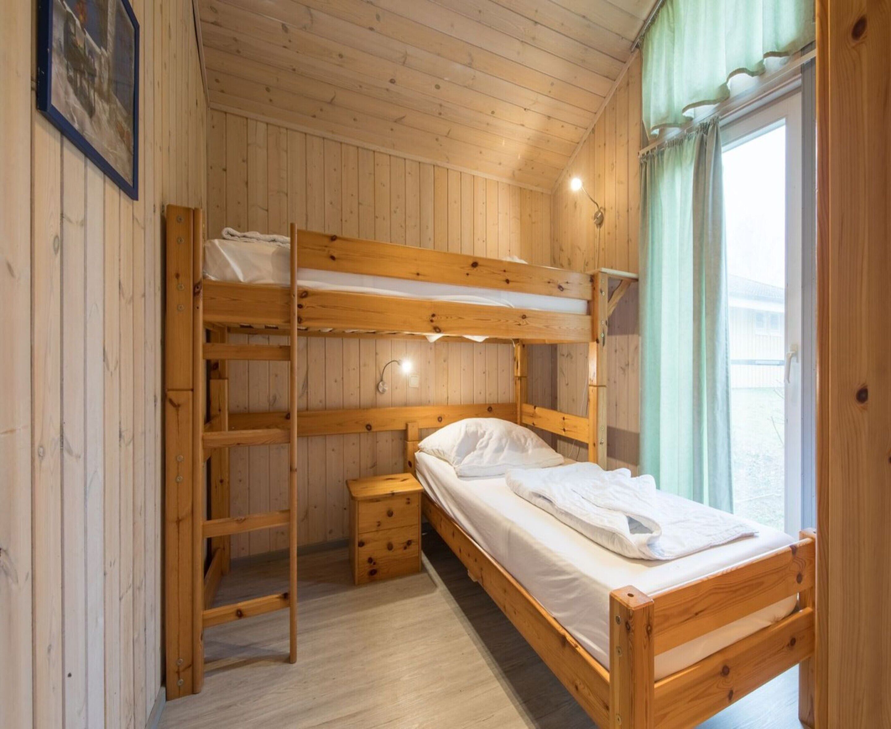 A bunk bed room with a wooden floor, a small wooden dresser, and a window with curtains.