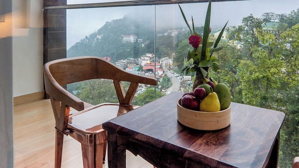 A balcony with a wooden table and chair, a bowl of fruit, and a view of a town through a glass railing. - Xyst Notting Hill (Dehradun)