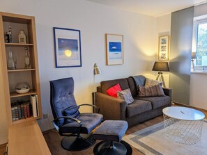 A living room with a sofa, armchair, and a small round table. There are framed pictures on the wall, a lamp, and a bookshelf with books and decorative items.