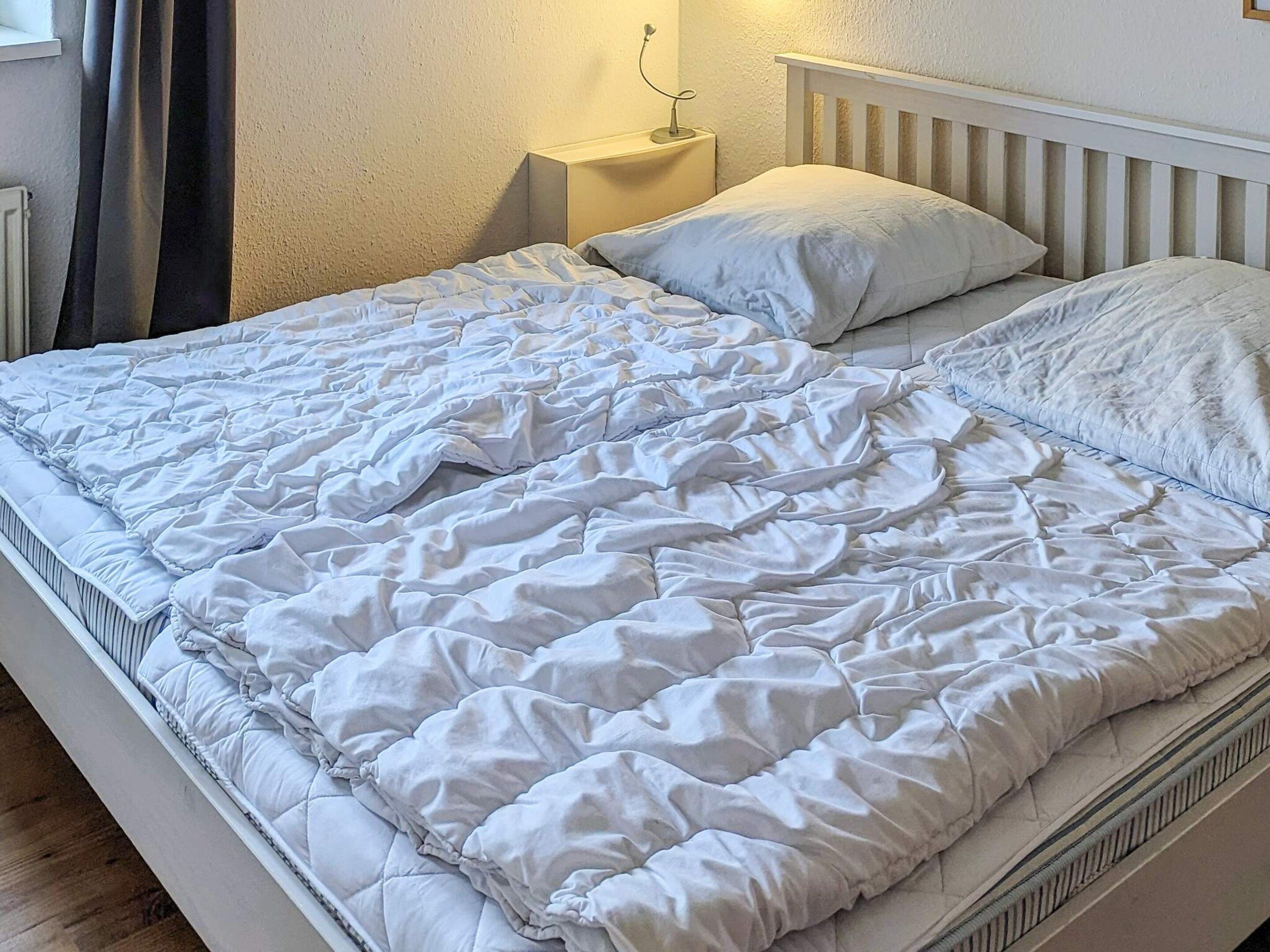 Uma cama com colchão e travesseiros acolchoados em branco, uma mesinha de cabeceira com abajur e cabeceira branca.