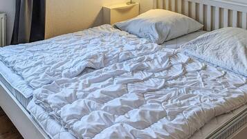 Uma cama com colchão e travesseiros acolchoados em branco, uma mesinha de cabeceira com abajur e cabeceira branca.