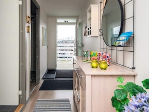 Un pasillo con piso de madera, felpudo negro, una alfombra pequeña, un mueble de madera con espejo y una planta.