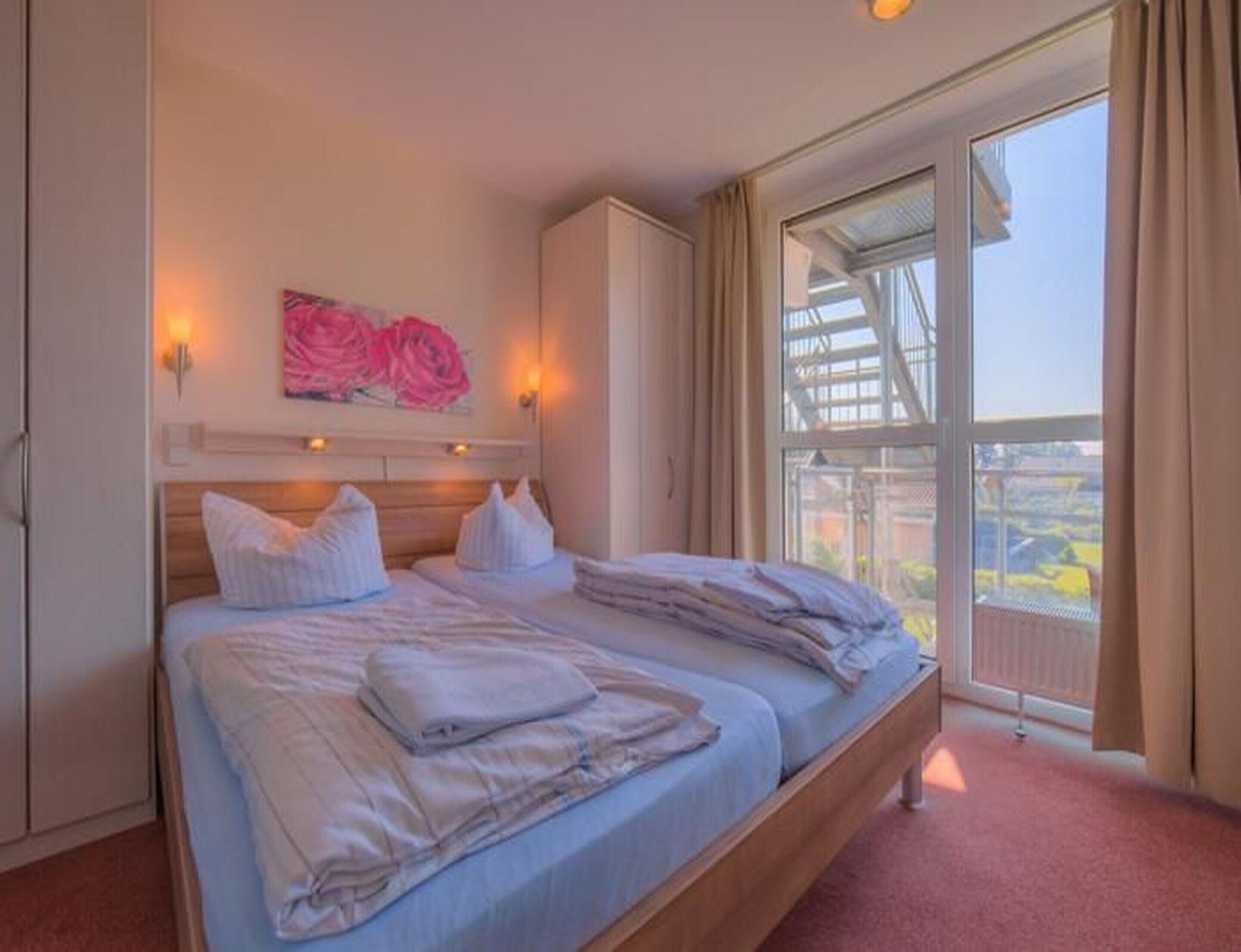 A double bed with a wooden headboard, a painting of pink flowers, and a view of a balcony with a railing.