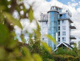 Un edificio moderno de varios pisos con una torre cilíndrica de vidrio, rodeado de vegetación.