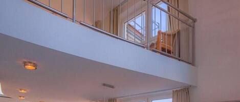 A spacious living area with a dining table, chairs, and a sofa. There is a balcony with a railing and a view of the outside.