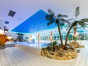 Un espace piscine intérieur moderne, doté d’un plafond en verre, de palmiers et d’un sol carrelé en pierre.