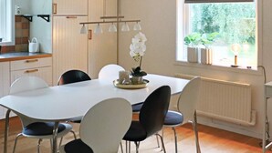 Un comedor con una mesa blanca, cuatro sillas, un jarrón con flores blancas y un ventanal con vistas a zonas verdes.
