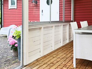 A covered porch with a white bench and a table, a red building in the background, and a potted plant with pink flowers. - 4 Star Holiday Home in Mellbystrand (Mellbystrand)