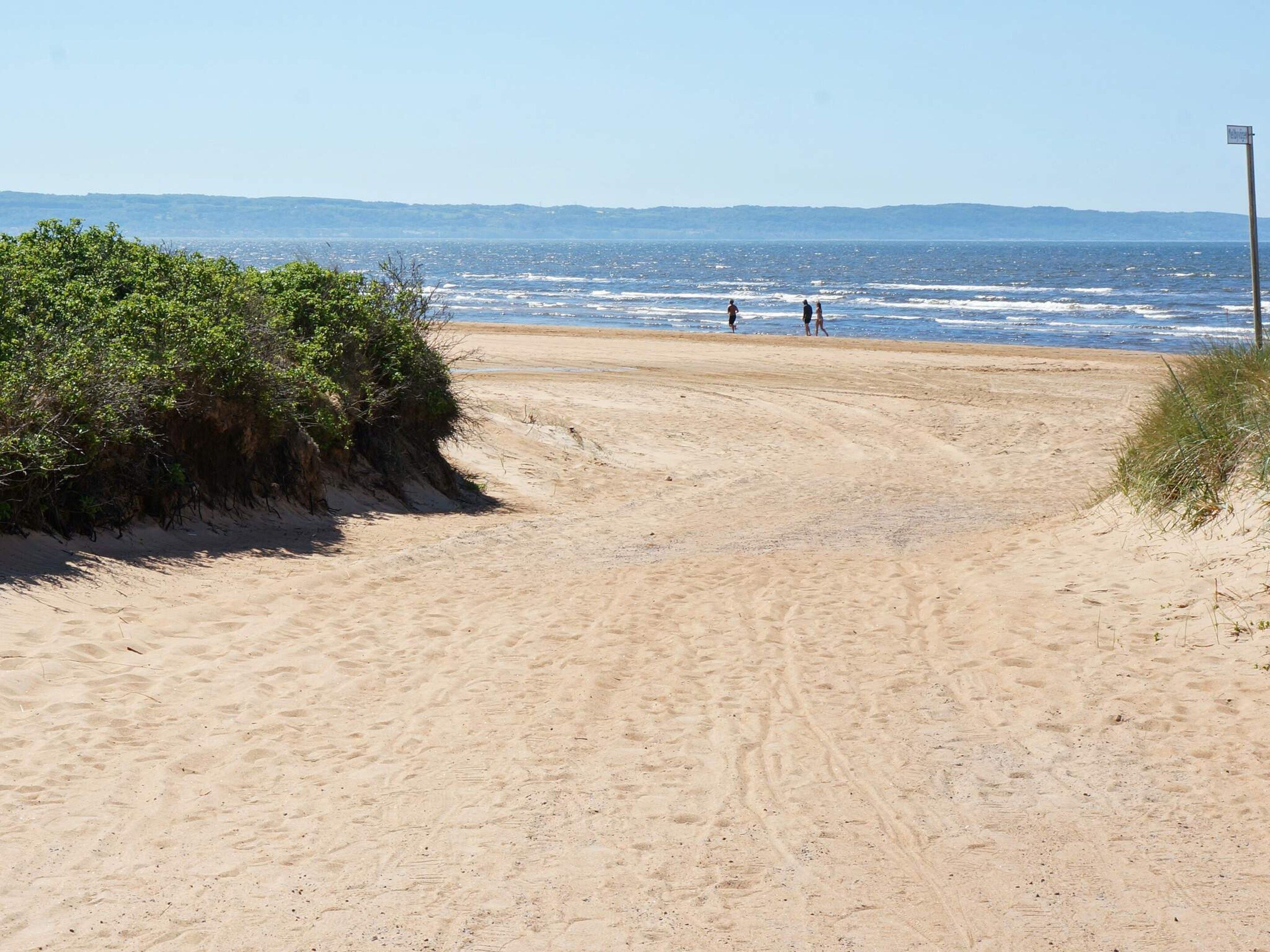 A sandy beach path leading to the ocean with a clear sky and distant hills.