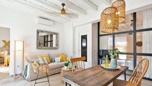 Ein geräumiges Wohnzimmer mit einem Holzessttisch, Korbstühlen, einer Couch mit Kissen, einem großen Spiegel und einem Deckenventilator.
