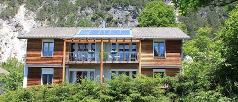 Une maison en bois à deux étages, dotée d’un balcon et entourée de verdure et de montagnes.