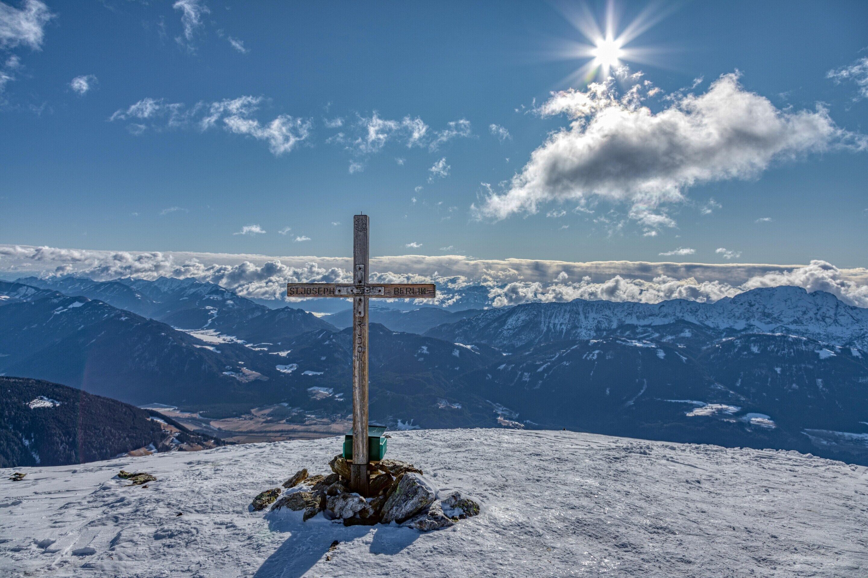Una cruz de madera con señales de dirección en la cima nevada de una montaña, rodeada de montañas cubiertas de nieve y un cielo despejado.