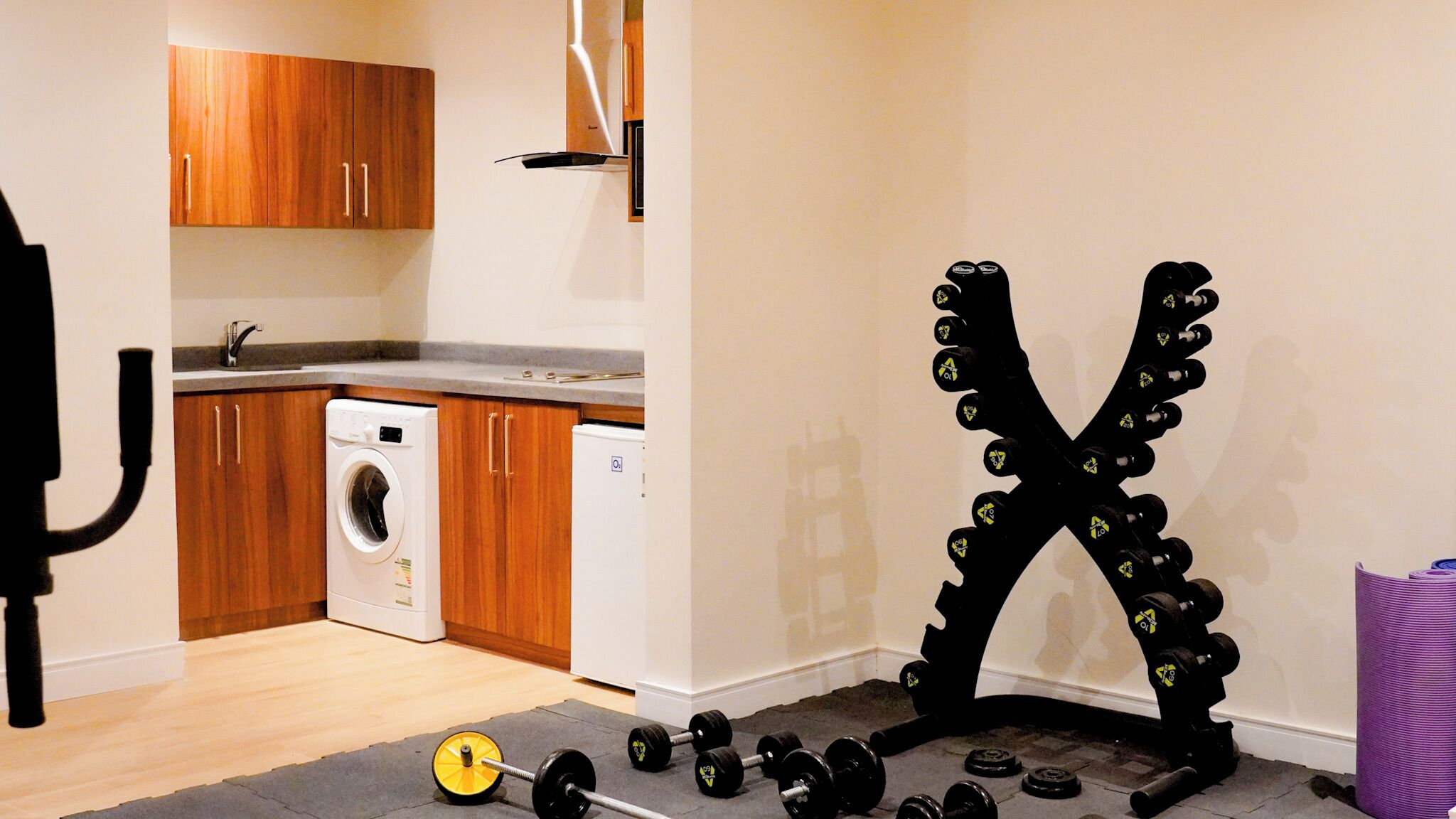Quarto com cozinha, máquina de lavar, suporte para halteres e tapete de yoga.