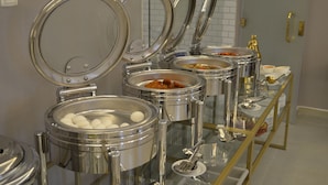 Een buffetopstelling met meerdere roestvrijstalen warmhoudschalen, waarvan sommige eten bevatten en andere witte, bolvormige items, waarschijnlijk eieren, gepresenteerd op een glazen toonbank met een gouden lijst.