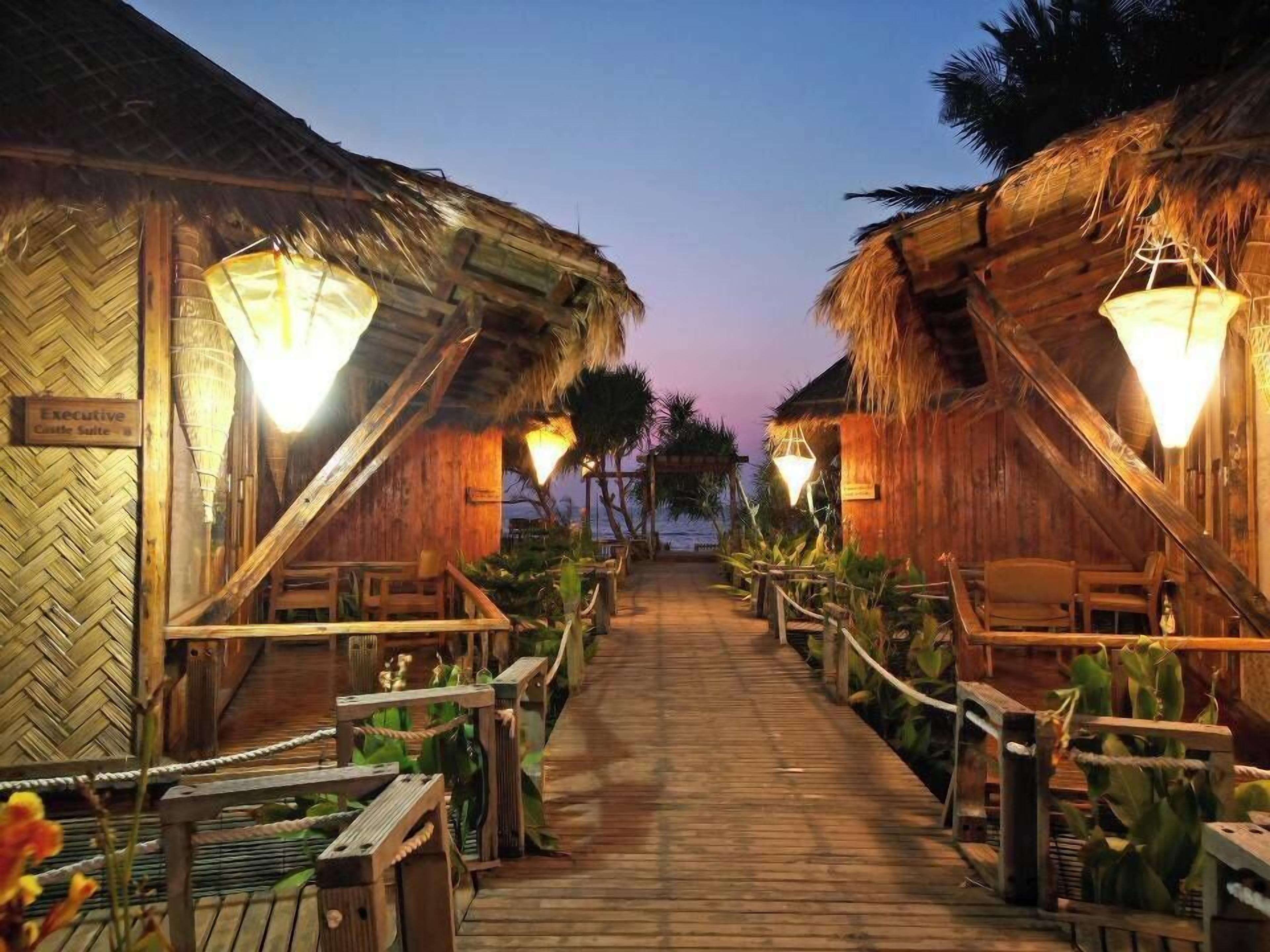 Een houten pad dat leidt naar rieten hutten met hangende lantaarns, omgeven door tropische planten.