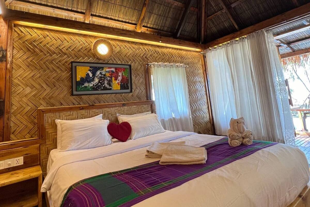 Habitación de hotel con pared de madera en patrón de espiga, cama con ropa blanca, almohada en forma de corazón rojo, cuadro abstracto enmarcado y ventana con cortinas blancas.
