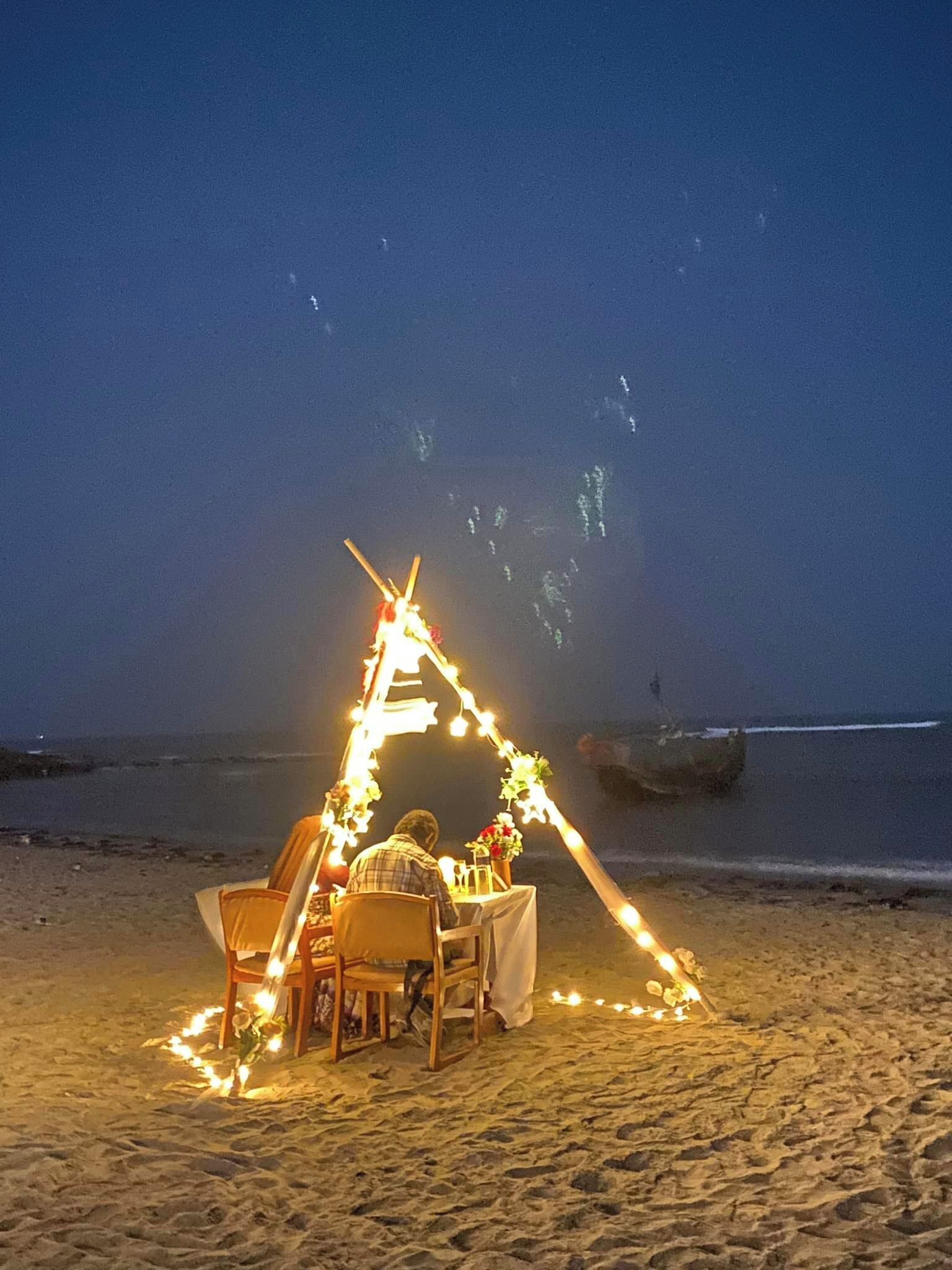 A beachside dining setup with a tent and chairs, decorated with lights and flowers, overlooking the ocean.