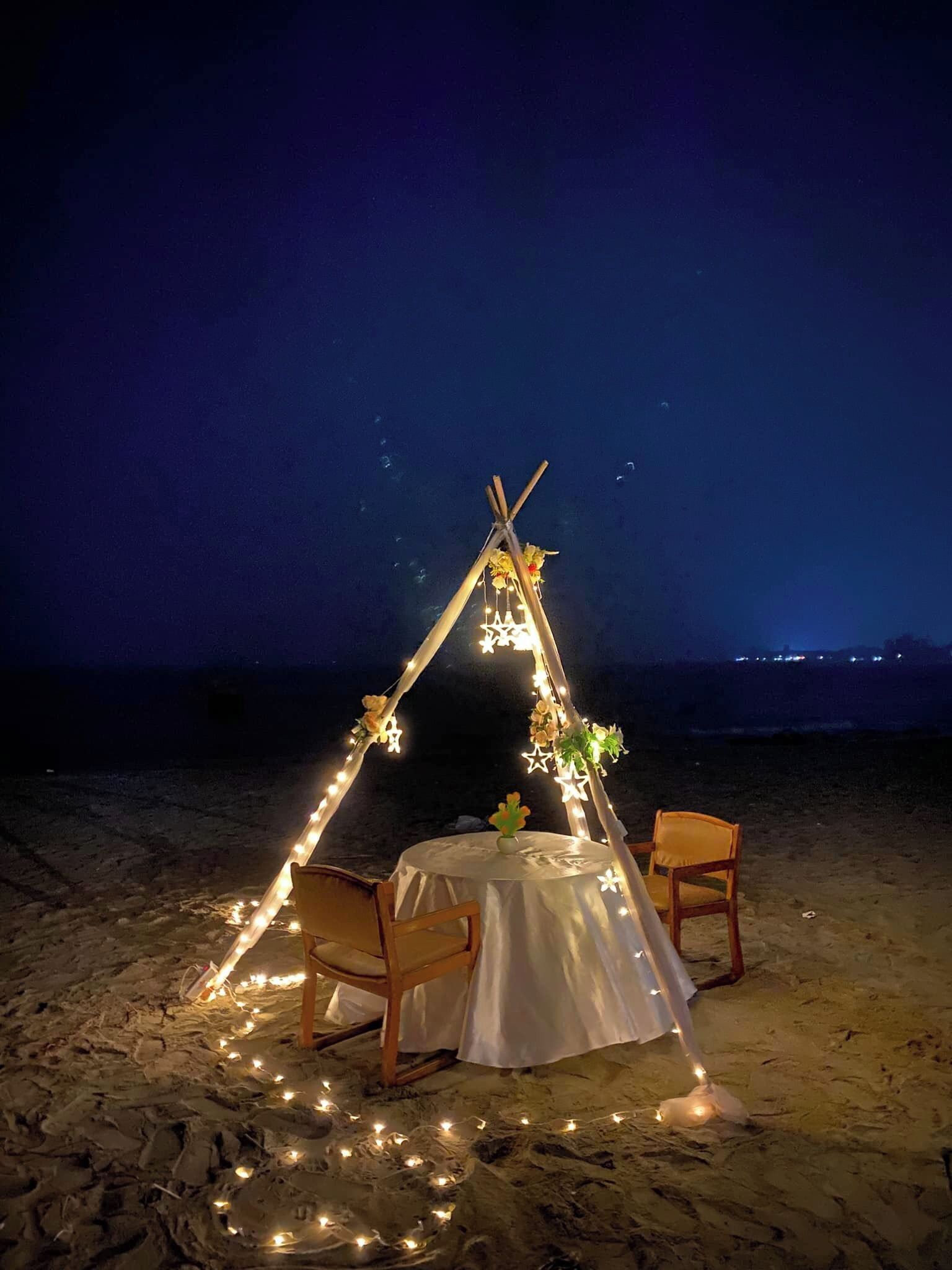 A tent set up on a beach with a table and chairs underneath, decorated with string lights and flowers.