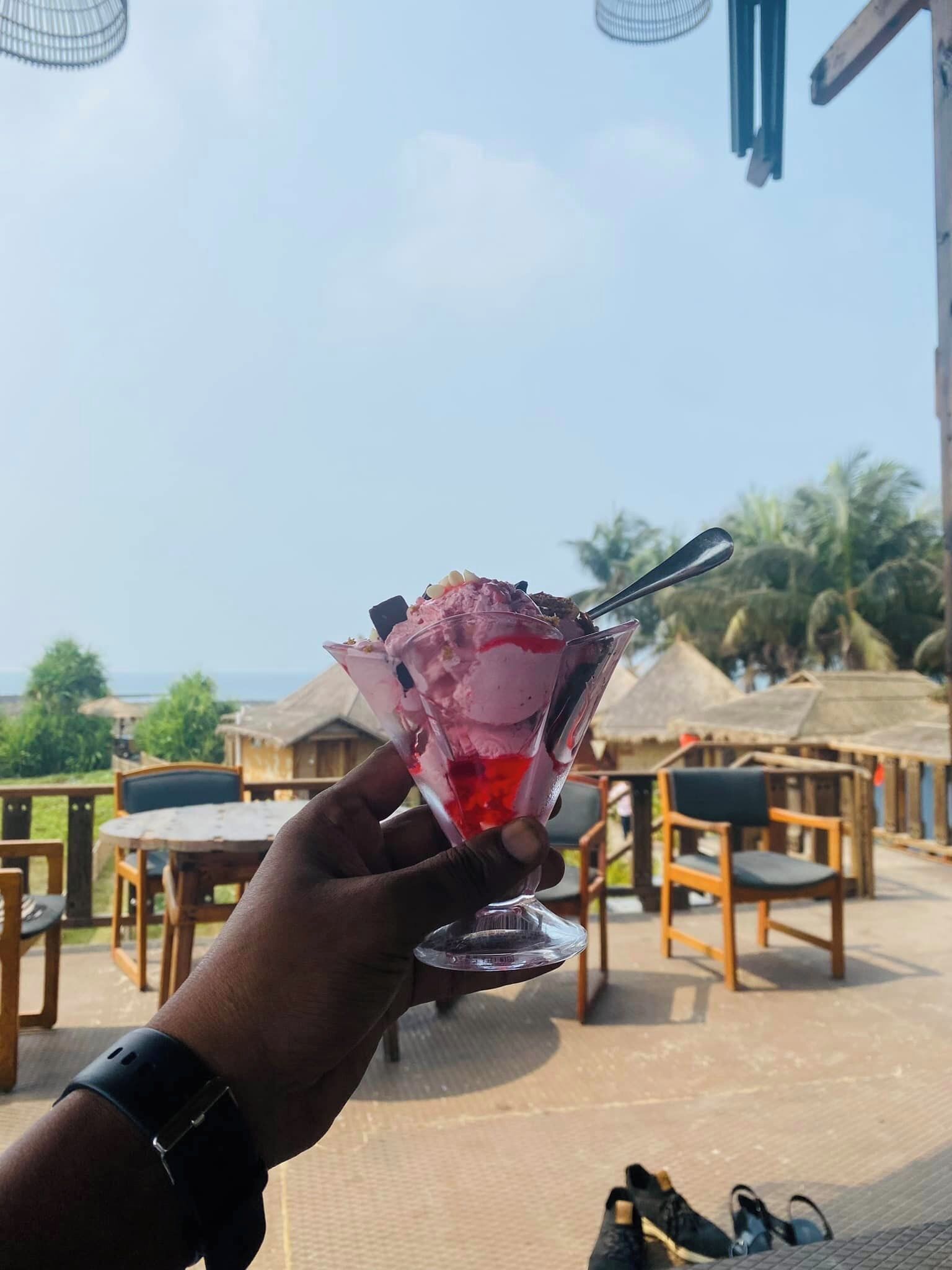 A hand holding a cocktail glass with a spoon, set against a backdrop of a beachside patio with wooden furniture and thatched umbrellas.