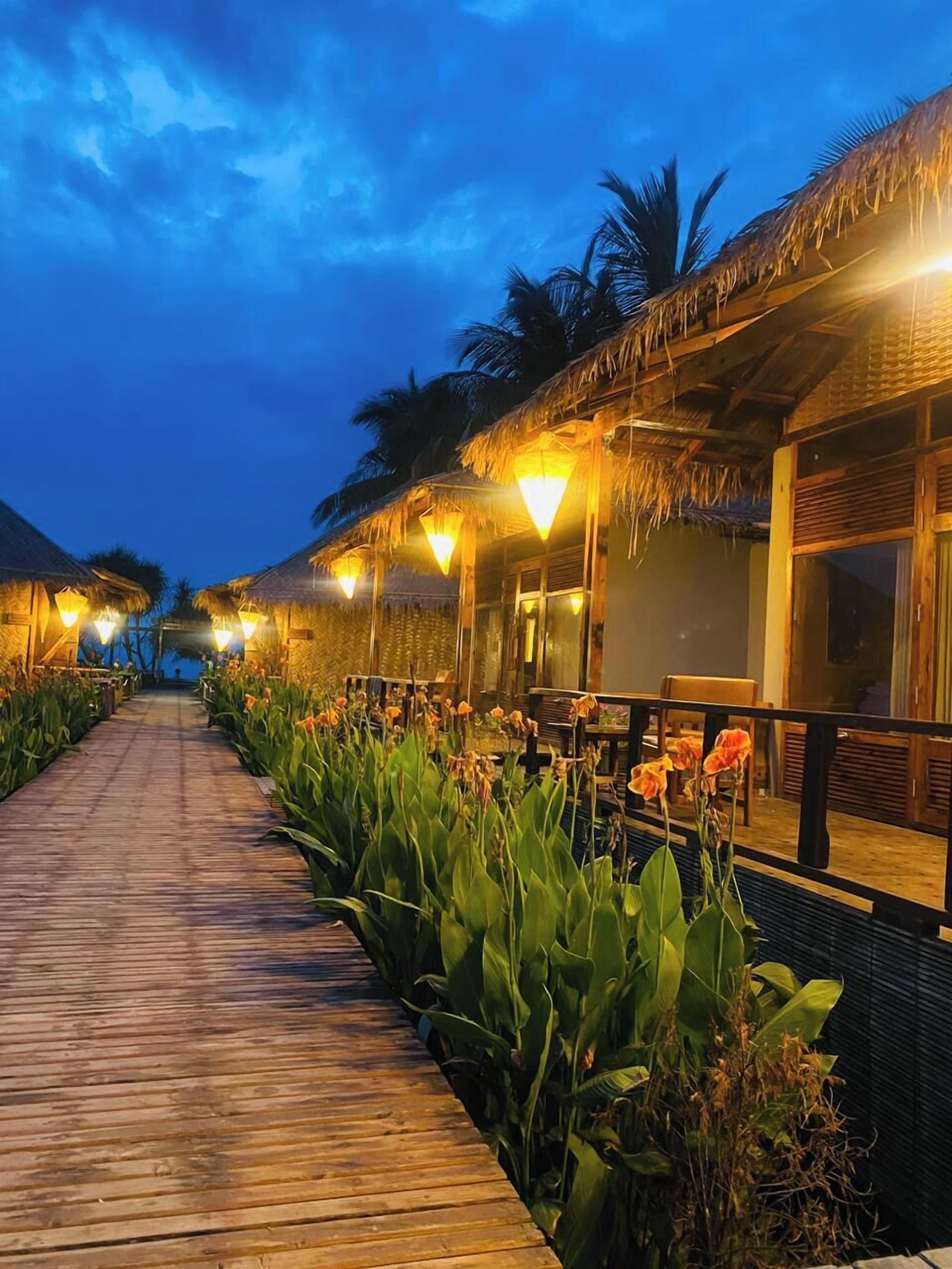 A wooden pathway leading to overwater bungalows with thatched roofs and warm lighting.