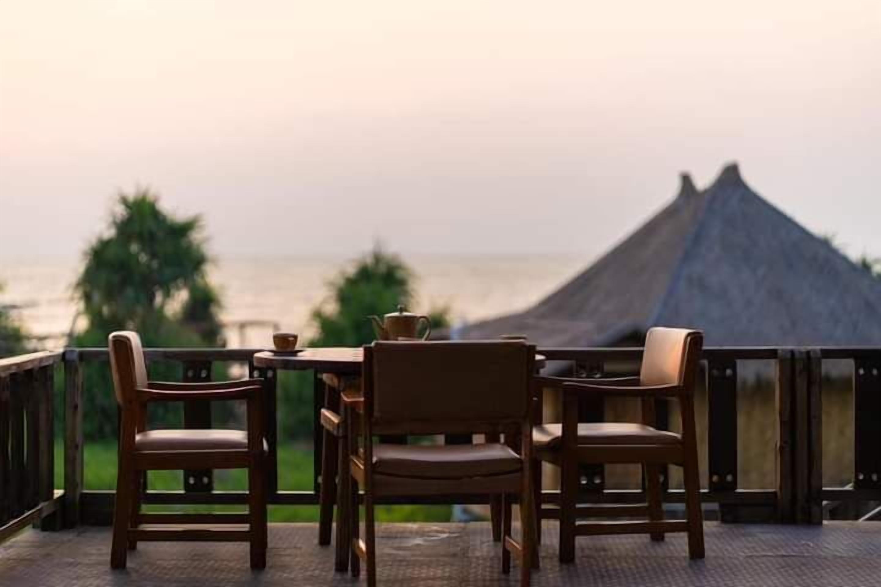 A terrace with wooden chairs and a table, overlooking a beach with a thatched-roof structure.