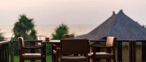 A terrace with wooden chairs and a table, overlooking a beach with a thatched-roof structure.