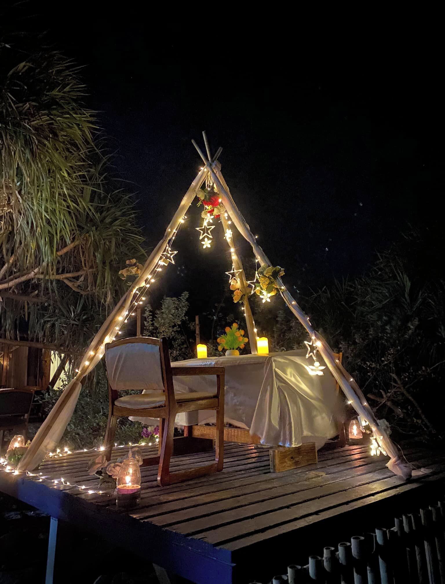 Cómodo comedor al aire libre con mesa y sillas de madera bajo un toldo triangular decorado con luces de cuerda y adornos de estrellas.