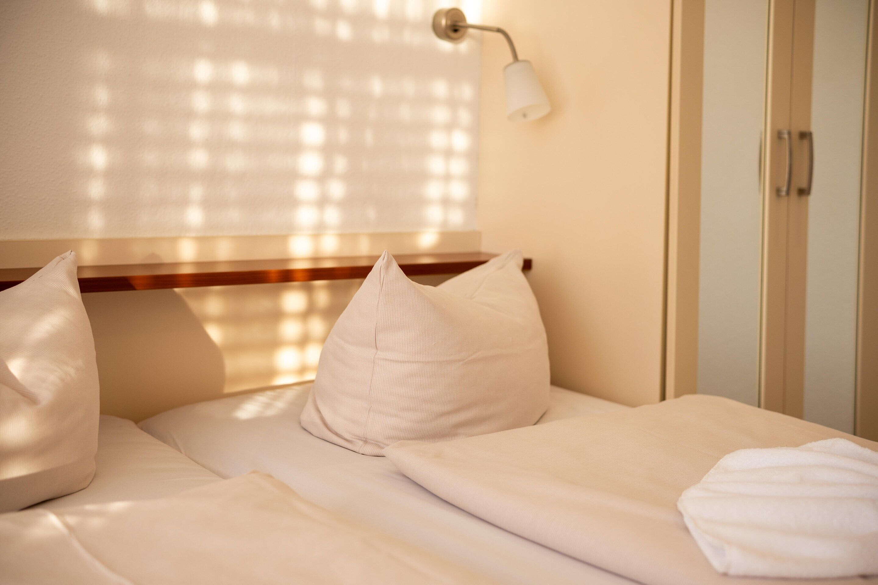 A neatly made bed with white linens and pillows, a bedside lamp, and a window with blinds.