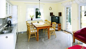 A dining area with a wooden table and chairs, a kitchen with white cabinets, a television, and a red sofa. - Doppelhaushalften Boltenhagen in Deutschland (Boltenhagen)