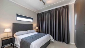 Ein modernes Schlafzimmer mit einem großen Bett, zwei Nachttischen inklusive Lampen, einem Fenster mit Jalousien und einem dunklen Vorhang.