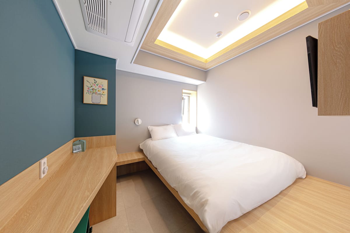 A modern bedroom with a bed, a wooden desk, a wall-mounted TV, and a ceiling light fixture.