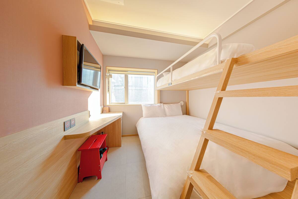 A bunk bed room with a TV mounted on the wall, a window, and a red bench.