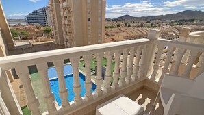 En balkon med hvide gelændere, udsigt over en swimmingpool og et boligområde med bygninger samt et kuperet landskab.