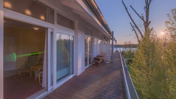 Una terraza de madera con mobiliario exterior, ventanas amplias y vistas a una masa de agua.