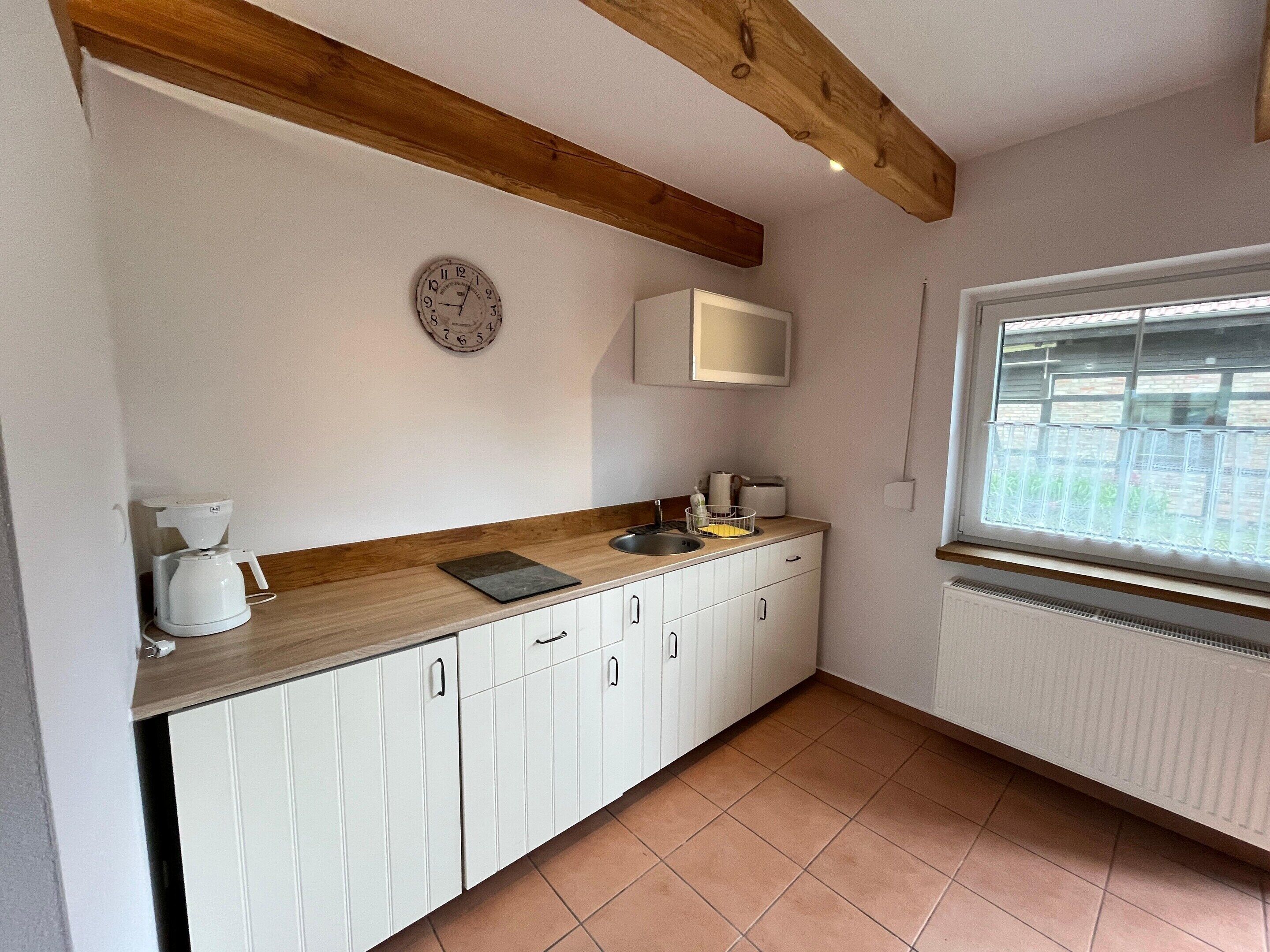 A small kitchen with white cabinets, a wooden countertop, a coffee maker, a sink, a window with a view, and a clock on the wall.