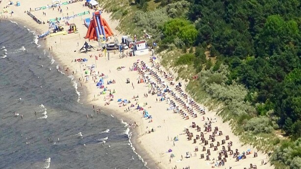 Vista aérea de una playa con filas de sillas y sombrillas, un tobogán inflable grande y abundante vegetación a la derecha.