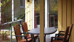 Ein überdachter Außenbereich mit Tisch und Stühlen, einer Holzterrasse und Blick auf ein Gebäude mit Ziegeldach.