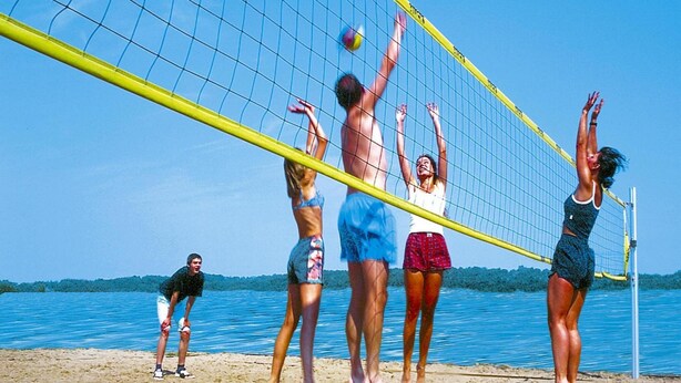 Eine Gruppe von Menschen spielt Beachvolleyball an einem sandigen Strand, im Hintergrund ein klarer, blauer Himmel und Wasser.