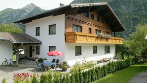 Un bâtiment à deux étages doté d’un balcon, d’une terrasse couverte avec des chaises et un parasol, et ayant une chaîne de montagnes en toile de fond.