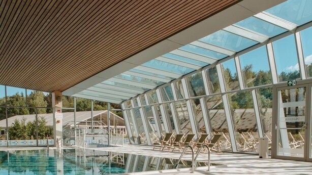 An indoor swimming pool with clear water, wooden ceiling, and glass walls offering views of the outdoors. - The Brookside Escape (Antoing)