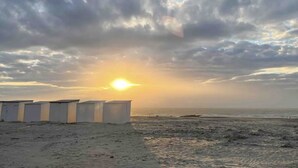 A beach with a row of white buildings, possibly a hotel, under a cloudy sky with the sun setting or rising. - Cosy Apartment With Terrace Near Beach (Nieuwpoort)