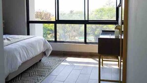 A bedroom with a large window, a bed, a desk, and a patterned rug. - Stylish & Elegant With Epic Views Near Menlyn (Pretoria)