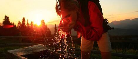 Une personne boit de l’eau à un robinet en métal, à l’extérieur, au coucher du soleil.