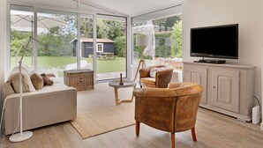 A modern living room with a sofa, armchairs, a television, and a view of a garden through large glass doors. - Holiday Home in Kamperland Near Beaches (Kamperland)