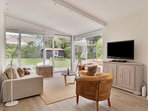 A modern living room with a sofa, armchairs, a television, and a view of a garden through large glass doors. - Holiday Home in Kamperland Near Beaches (Kamperland)