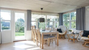 Une pièce spacieuse avec une table à manger en bois, des chaises et une petite table ronde avec un siège. De grandes fenêtres laissent entrer beaucoup de lumière naturelle et offrent une vue sur l’extérieur.